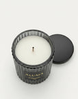 An open black ribbed glass candle jar with white wax and a single wick, shown on a light gray background with its black lid beside it. The jar is labeled ALUMA.
