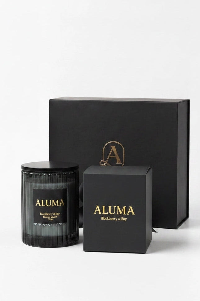 A black glass jar candle with a black lid and gold lettering, displayed alongside its black gift box and a smaller black presentation box with gold text. The brand name 'ALUMA' is clearly visible on the labels.