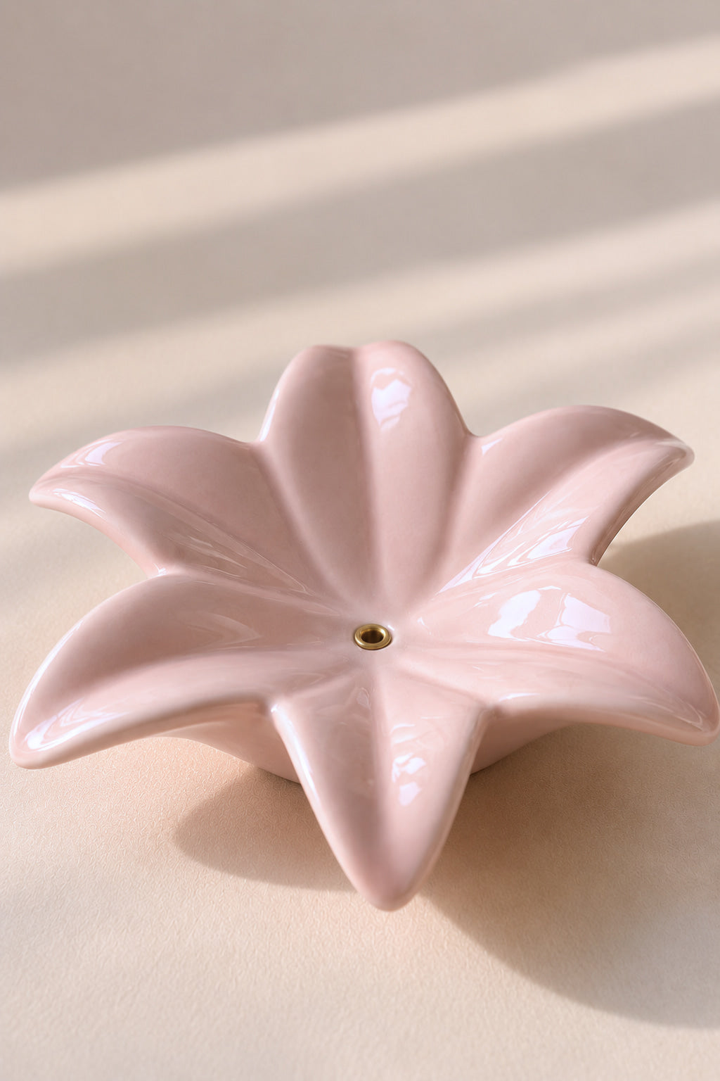A glossy, five-petal flower-shaped ceramic dish in a soft light pink color, featuring a small gold accent at the center, set against a warm beige background with soft diagonal shadows.
