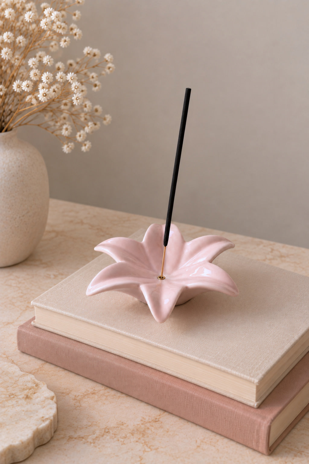 A pink ceramic lotus-shaped incense holder sits on stacked books, with a black incense stick inserted and dried flowers in a vase in the background.