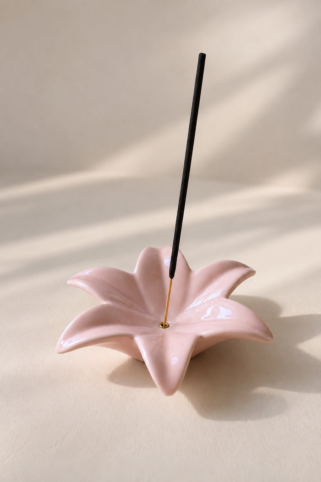 A glossy light pink flower-shaped incense holder with a single black incense stick standing upright in the center, photographed on a warm beige surface with soft shadows.