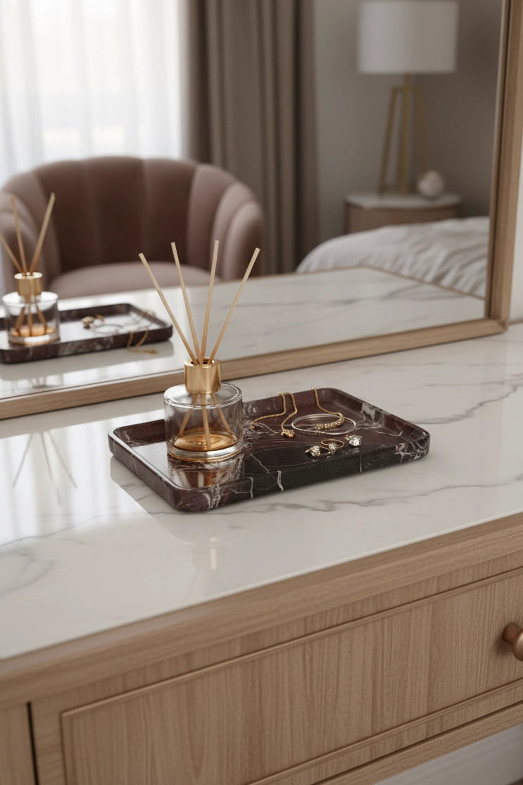 A modern vanity or dressing table with a light wood grain cabinet, a white marble-look top, and a large mirror. A dark marble tray holding a reed diffuser and small jewelry items sits on the surface, with the diffuser reflected in the mirror.