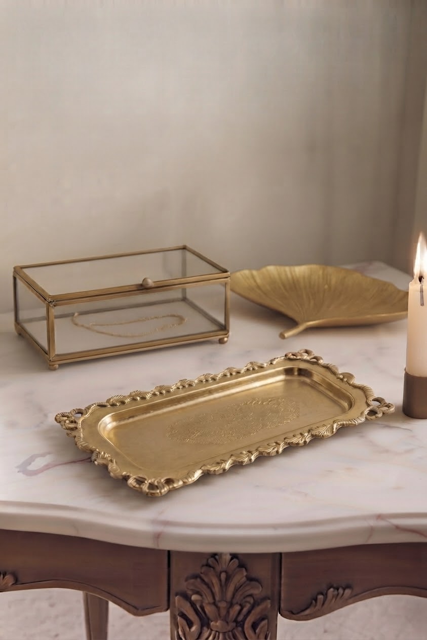 A set of decorative brass trays displayed on a white marble table, including a rectangular tray with ornate scalloped edges, a leaf-shaped tray, and a glass box with a brass frame.