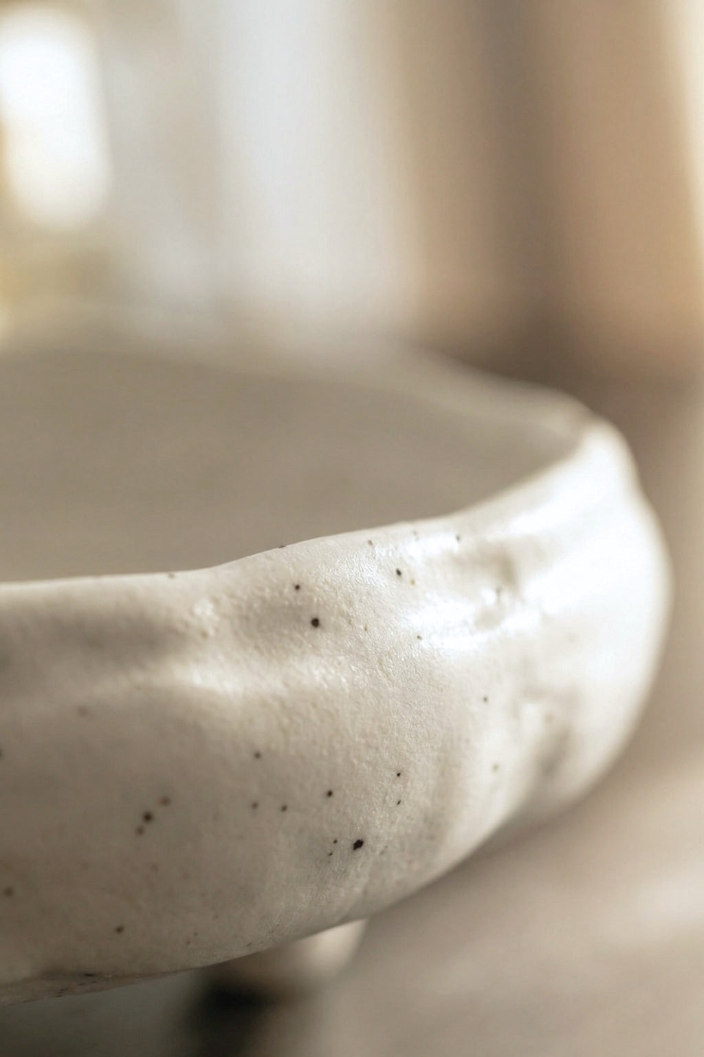 A close-up of a handmade ceramic bowl with a textured, organic shape and small black speckles, shown in a soft-focus warm-toned setting.