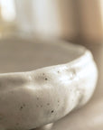 A close-up of a handmade ceramic bowl with a textured, organic shape and small black speckles, shown in a soft-focus warm-toned setting.