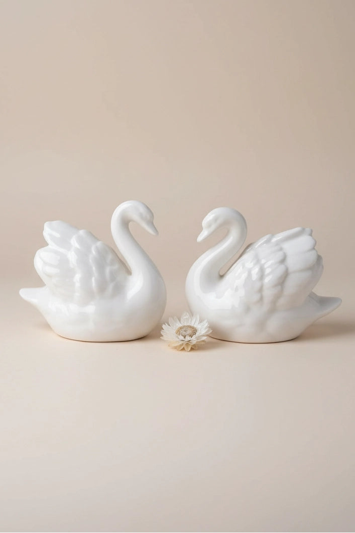 Two white ceramic swan figurines facing each other, with a small white flower placed between them, set against a solid beige background.