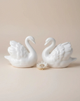 Two white ceramic swan figurines facing each other, with a small white flower placed between them, set against a solid beige background.