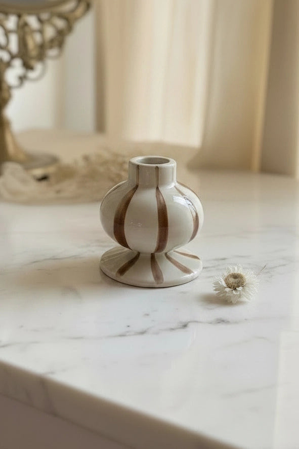 A small white ceramic vase with brown vertical stripes sits on a white marble tabletop in a softly lit room. A decorative mirror and cream curtains are blurred in the background.