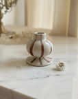 A small white ceramic vase with brown vertical stripes sits on a white marble tabletop in a softly lit room. A decorative mirror and cream curtains are blurred in the background.