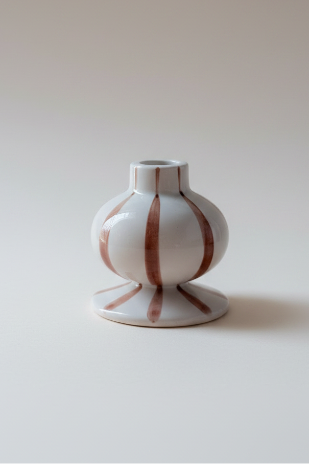 A small white ceramic bud vase with vertical terracotta-brown stripes, standing on a matching flared base against a warm beige gradient background.
