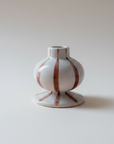 A small white ceramic bud vase with vertical terracotta-brown stripes, standing on a matching flared base against a warm beige gradient background.