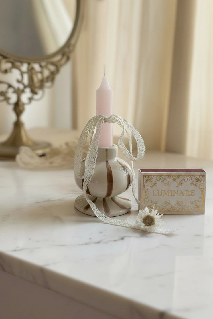 A decorative white ceramic candle holder with a pale pink taper candle, tied with a sheer white ribbon, displayed on a white marble tabletop next to a small matchbox.