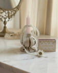 A decorative white ceramic candle holder with a pale pink taper candle, tied with a sheer white ribbon, displayed on a white marble tabletop next to a small matchbox.