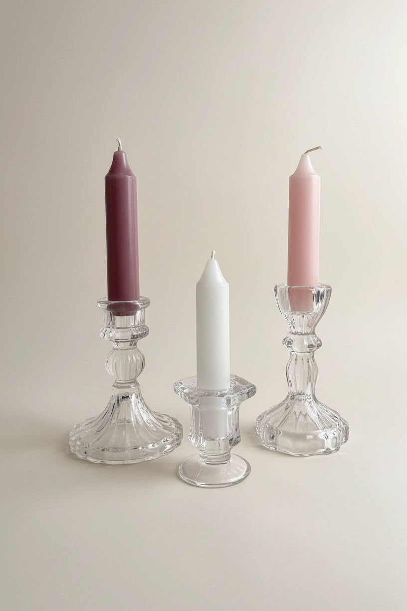 Three taper candles in clear glass candlesticks on a warm beige background: one dusty mauve, one white, and one soft pink.