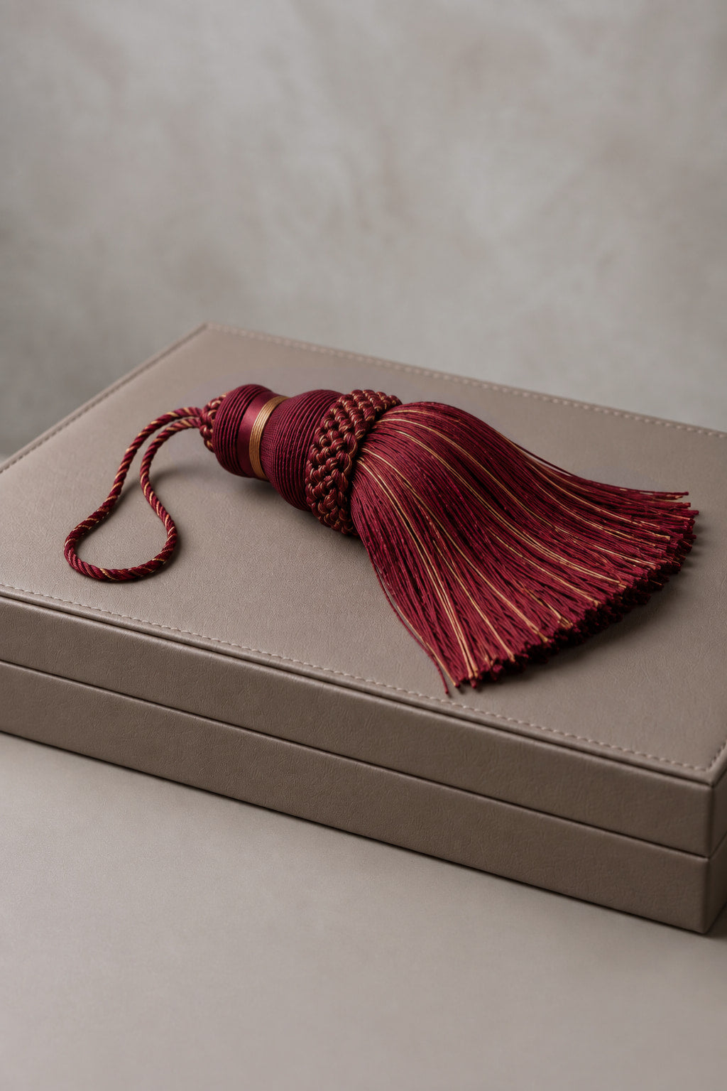 A decorative tassel with deep burgundy and gold striped threads, featuring a braided top and a matching cord loop, resting on a textured taupe leather box.