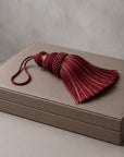 A decorative tassel with deep burgundy and gold striped threads, featuring a braided top and a matching cord loop, resting on a textured taupe leather box.