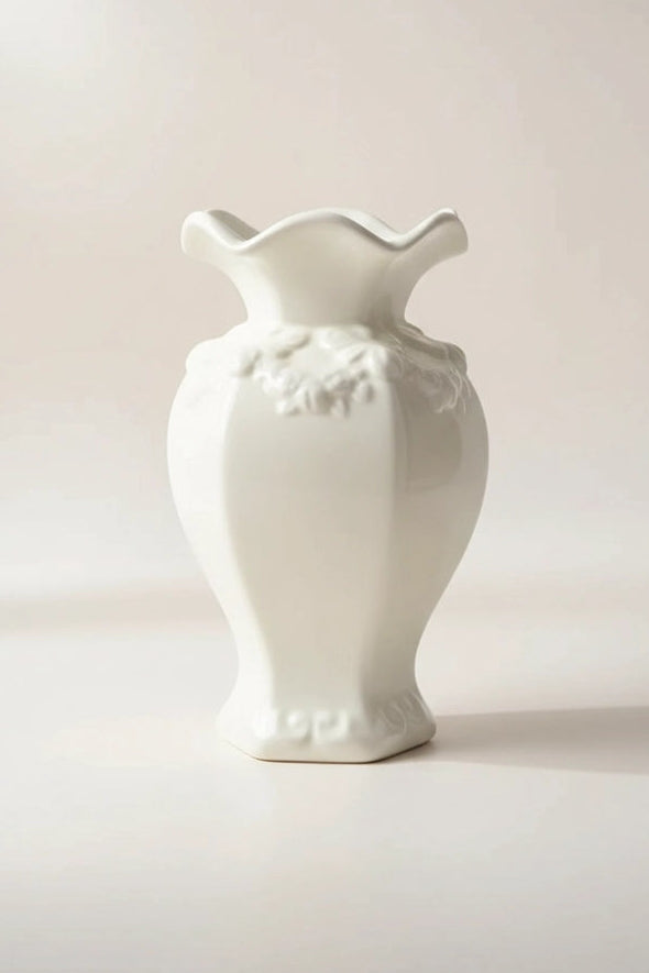 A  white ceramic vase with a flared, wavy rim and embossed floral detailing near the neck, photographed against a soft light beige studio background.