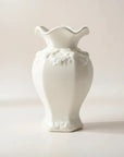 A  white ceramic vase with a flared, wavy rim and embossed floral detailing near the neck, photographed against a soft light beige studio background.