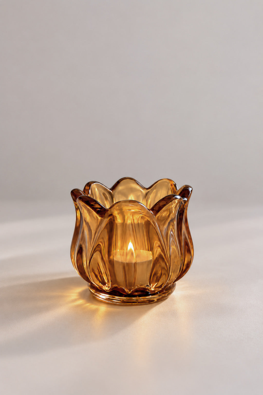 An amber glass candle holder with a fluted, lotus-like petal design, lit with a warm candlelight glow against a neutral light gray background.