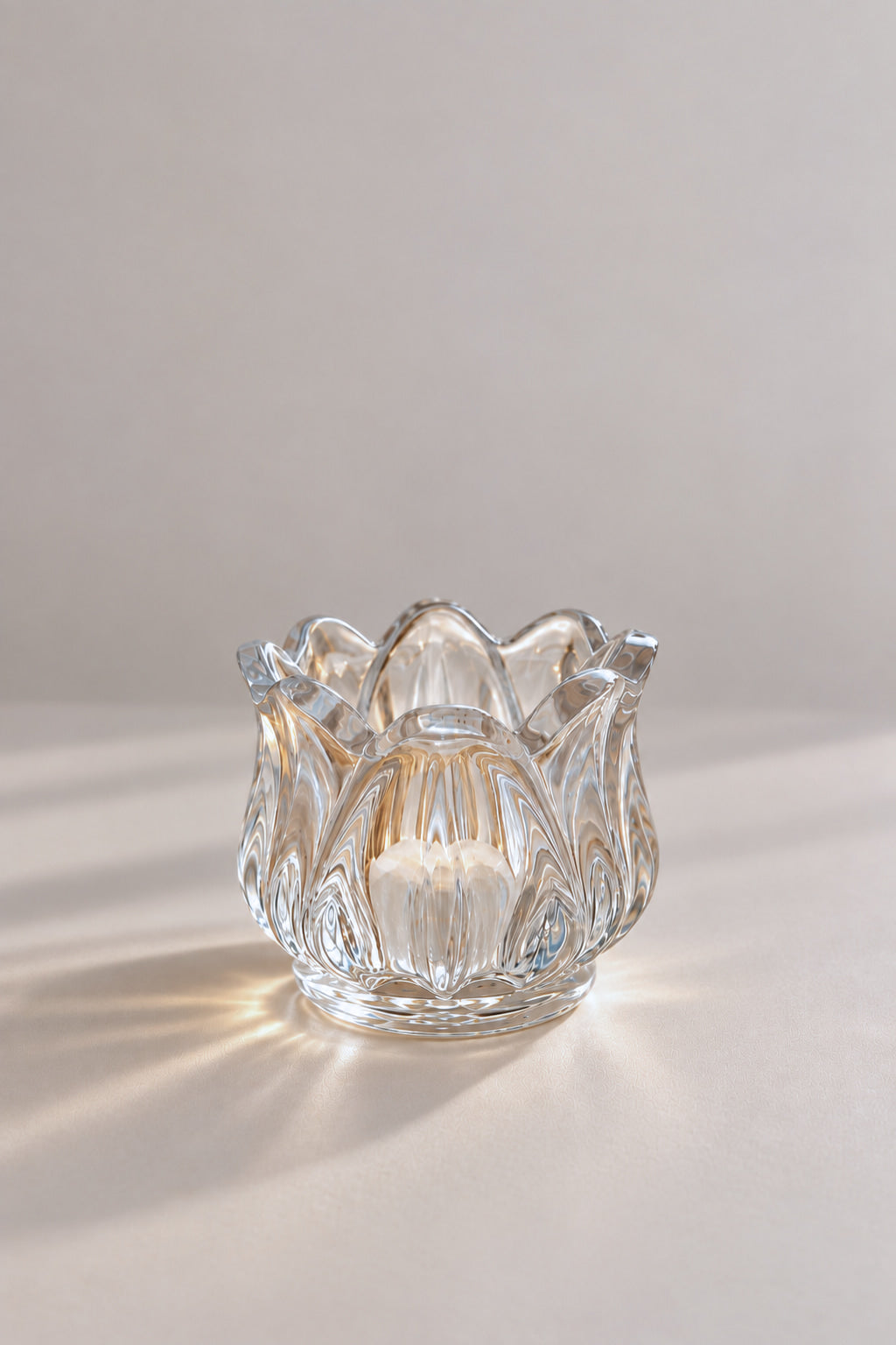 A clear glass candle holder with a lotus or lotus bud shape, featuring a faceted, textured pattern, set against a neutral light gray background with soft shadows.
