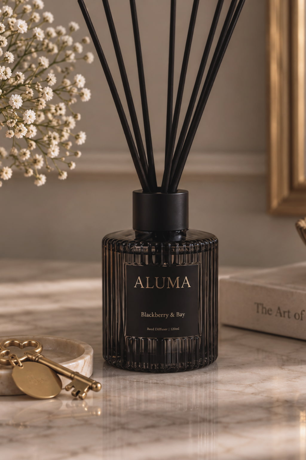 ALUMA Blackberry & Bay reed diffuser in a ribbed dark amber glass bottle with black reeds, styled on a marble surface with decorative keys, baby's breath, and a book.