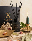 black aluma gift box, brass chamberstick, green lit candle, brass trinket dish, reed diffuser on a marble tray, dry flowers and bay leaves