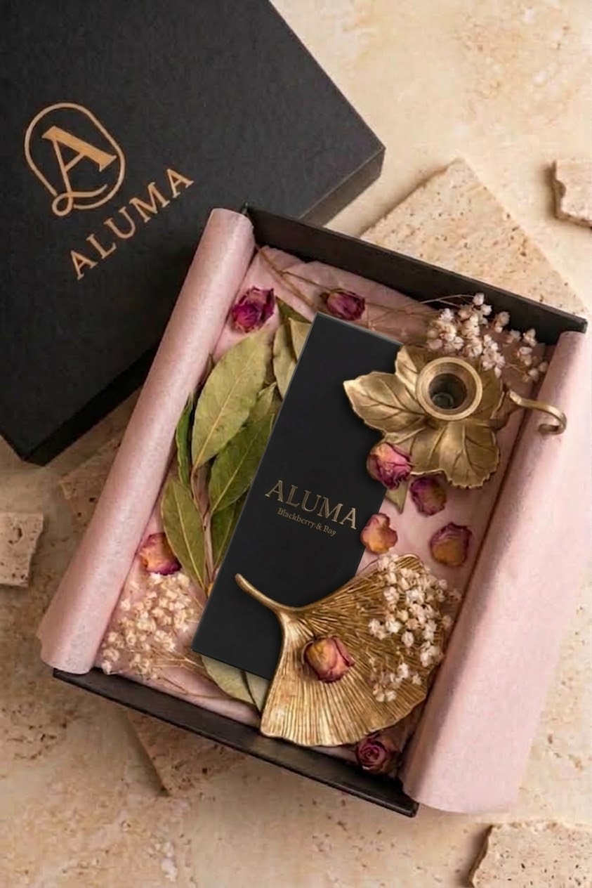 An open black gift box lined with blush pink tissue paper, styled with dried pink roses, green leaves, and gold-toned decorative leaves. A black card and a black box lid with the gold ALUMA logo are placed inside and beside the box on a beige stone surface.
