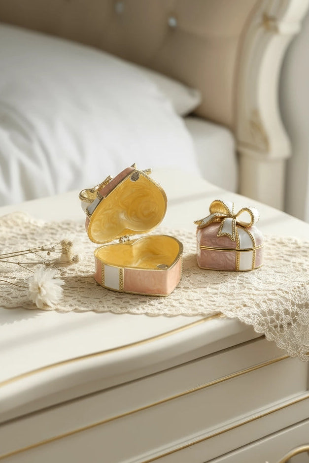 A decorative gold heart-shaped jewelry box and a small pink gift box with a bow, displayed on a cream-colored bedside table with lace in a bedroom setting.