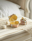 A decorative gold heart-shaped jewelry box and a small pink gift box with a bow, displayed on a cream-colored bedside table with lace in a bedroom setting.