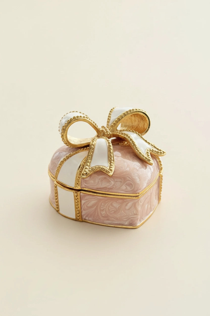 A small decorative gift box for rings  with a pink marbled body, white ribbon, and gold trim, topped with a large bow, photographed on a plain light beige background.