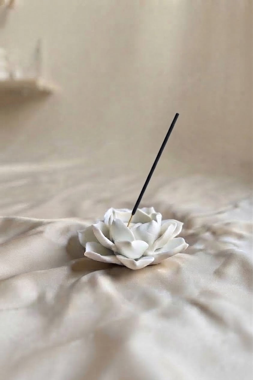 A white ceramic flower-shaped incense holder resting on draped beige fabric, with a single black incense stick inserted into its center.