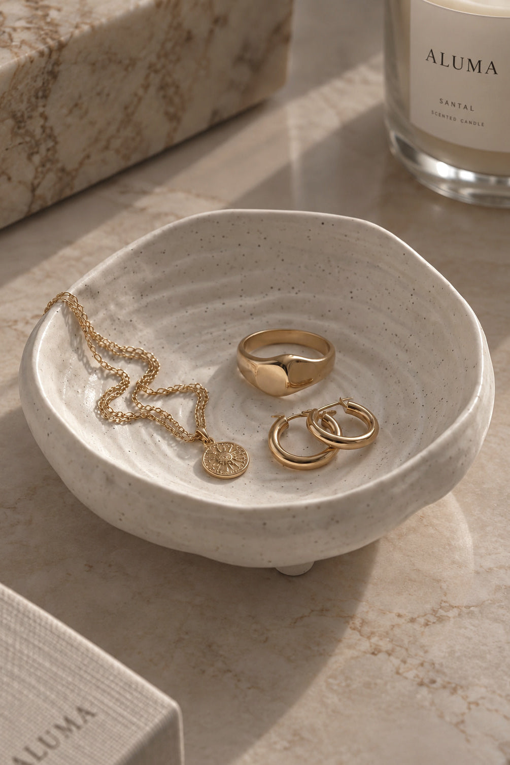 A small round ceramic jewelry tray with a textured speckled off-white finish, styled on a marble surface with gold rings and a necklace inside. An ALUMA candle and matching box are partially visible in the background.