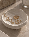 A small round ceramic jewelry tray with a textured speckled off-white finish, styled on a marble surface with gold rings and a necklace inside. An ALUMA candle and matching box are partially visible in the background.