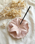 Pink ceramic incense holder with a black incense stick on a white fabric background