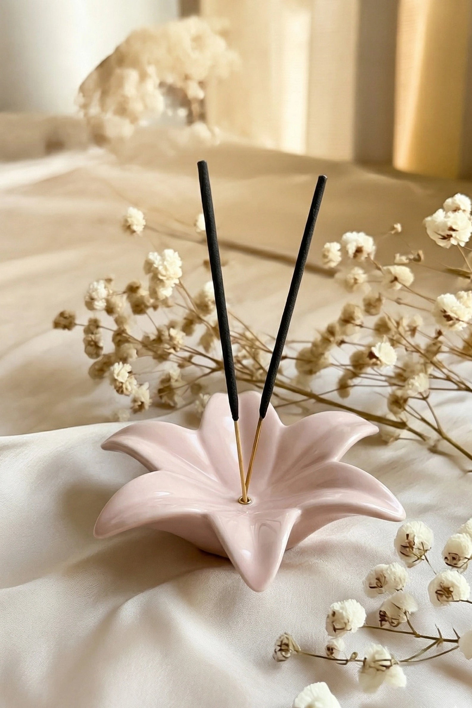 Pink ceramic incense holder with black sticks on a light fabric background with white flowers.