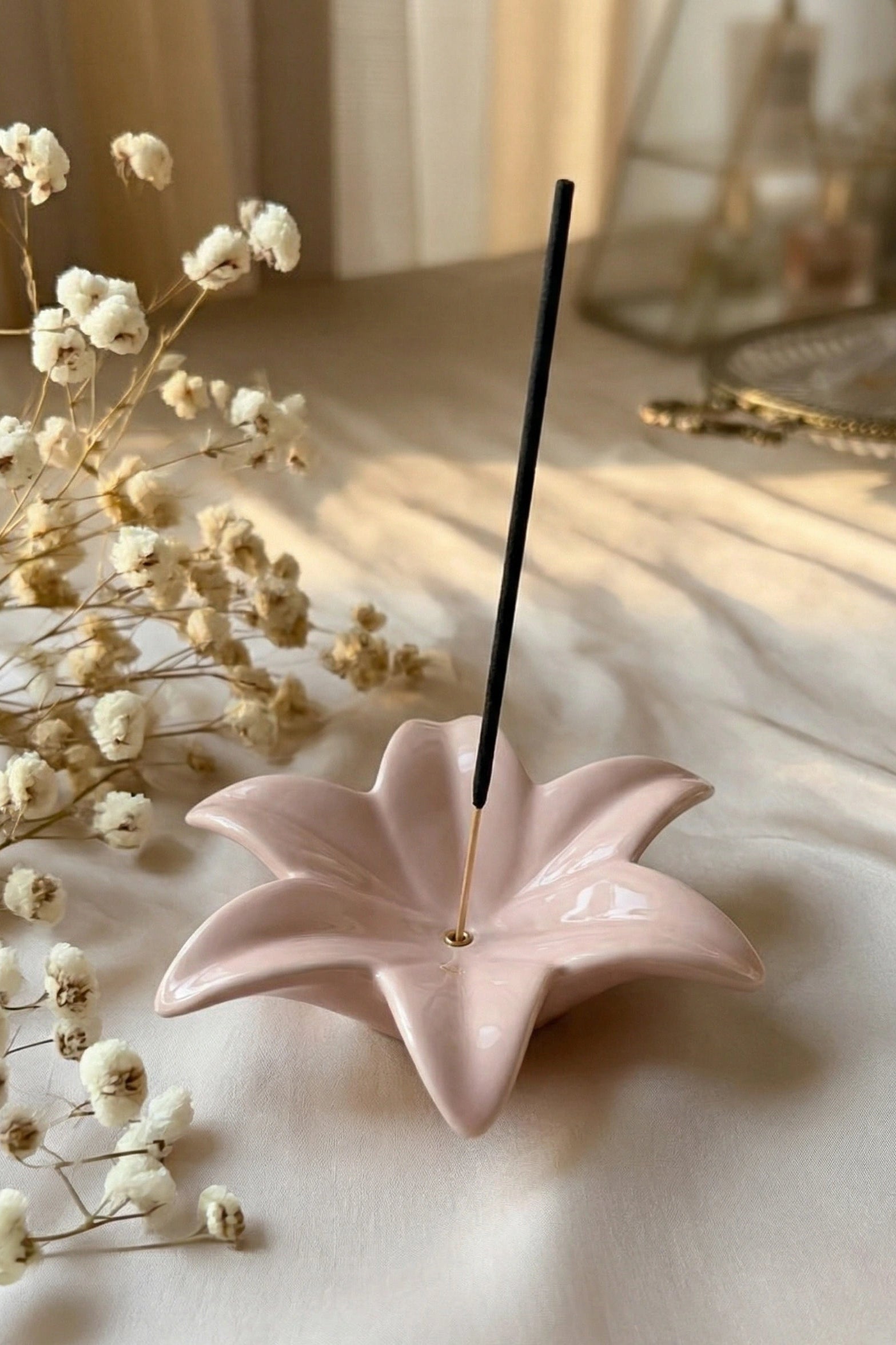 Pink lotus-shaped incense holder with a single black incense stick on a light surface with dried flowers.