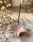 Pink lotus-shaped incense holder with a single black incense stick on a light surface with dried flowers.