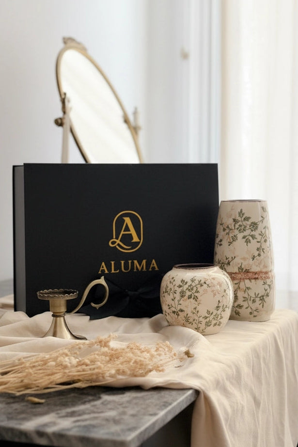 A black ALUMA gift box with gold lettering sits on a draped tablecloth beside ceramic vases and dried grasses in a softly lit interior setting.