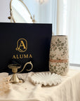 A styled interior scene showing a black ALUMA gift box with gold lettering, a cream floral ceramic vase, a small gold candle holder, and a white scalloped dish on a draped cream fabric surface in a bright room.