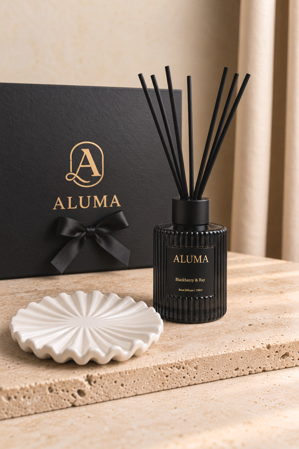 ALUMA reed diffuser bottle in black with black reeds, displayed on a white scalloped dish in front of a black gift box with gold ALUMA branding.