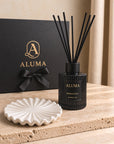 ALUMA reed diffuser bottle in black with black reeds, displayed on a white scalloped dish in front of a black gift box with gold ALUMA branding.