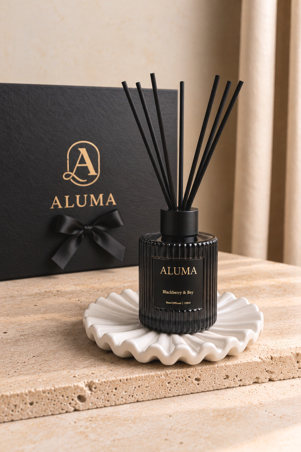 ALUMA reed diffuser bottle in black with black reeds, displayed on a white scalloped dish in front of a black gift box with gold ALUMA branding.