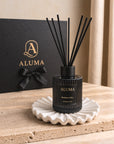 ALUMA reed diffuser bottle in black with black reeds, displayed on a white scalloped dish in front of a black gift box with gold ALUMA branding.