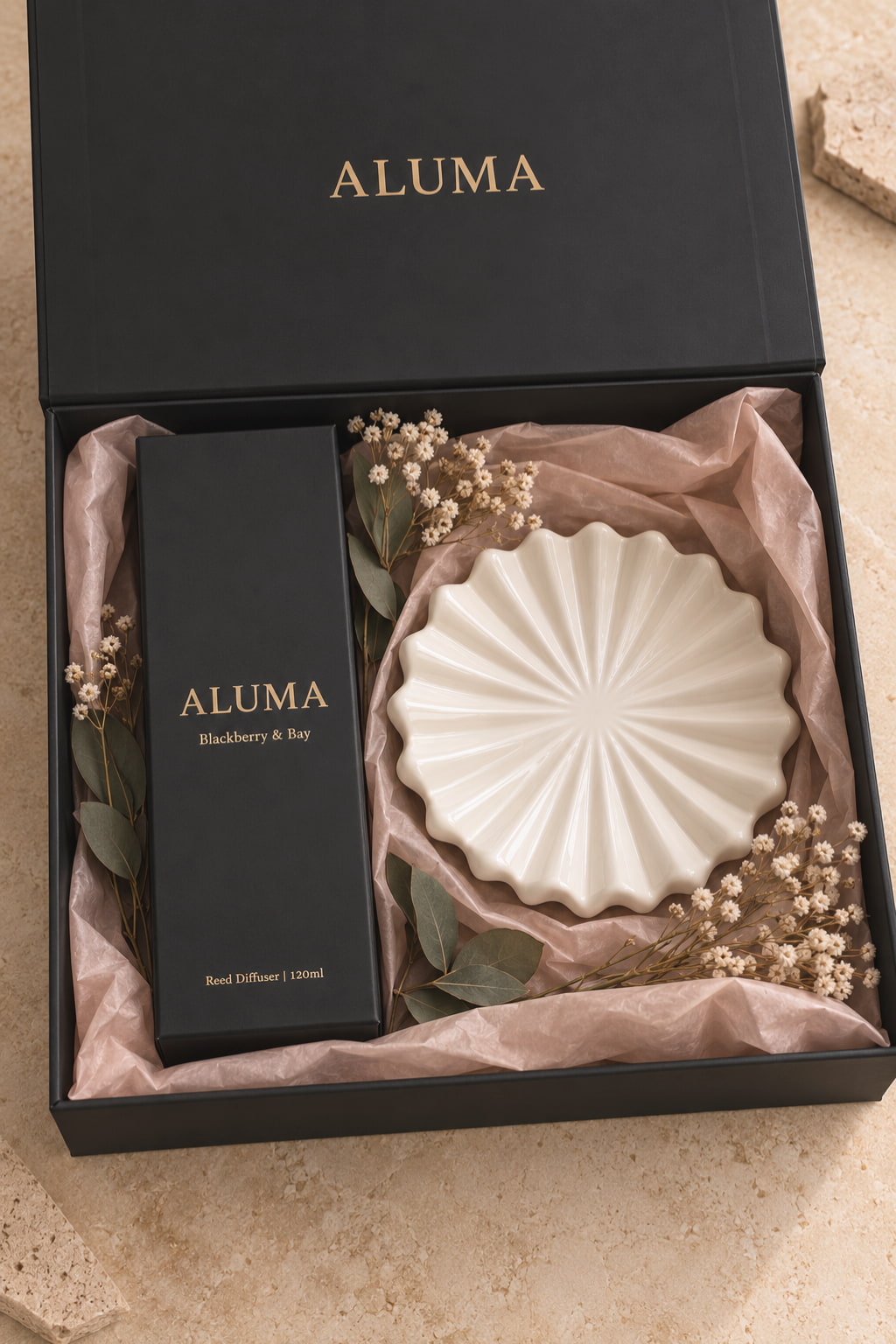 ALUMA Blackberry & Bay diffuser gift set in a black box with gold lettering, containing a black diffuser box and a white pleated round diffuser bowl on blush pink tissue paper with dried flowers.
