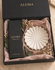 ALUMA Blackberry & Bay diffuser gift set in a black box with gold lettering, containing a black diffuser box and a white pleated round diffuser bowl on blush pink tissue paper with dried flowers.
