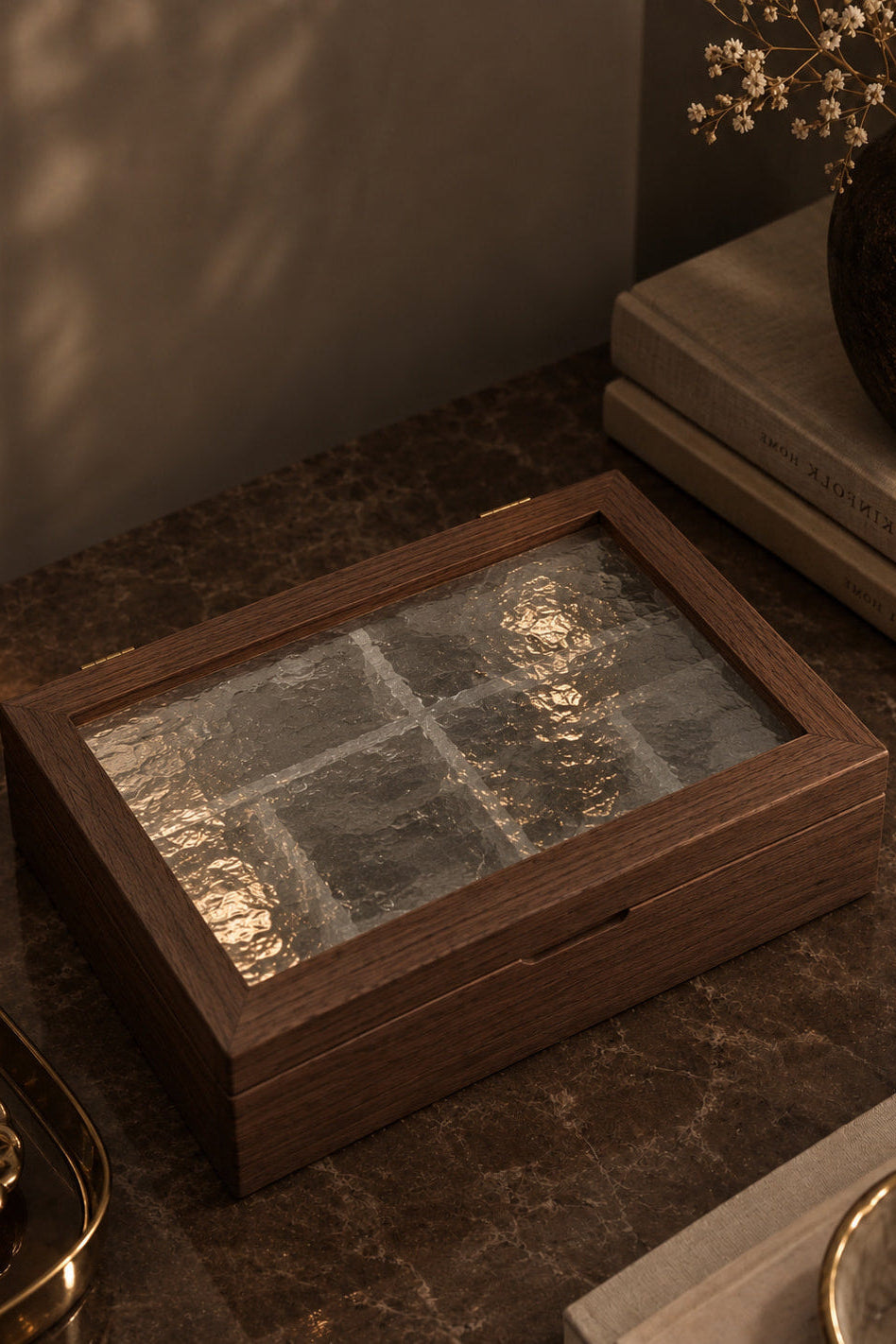 A rectangular wooden box with a textured glass lid, styled on a dark marble tabletop with books, a candle, and dried flowers in a warm, moody setting.