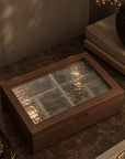 A rectangular wooden box with a textured glass lid, styled on a dark marble tabletop with books, a candle, and dried flowers in a warm, moody setting.