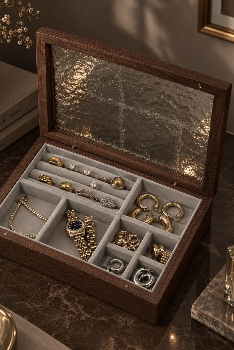 An open dark wood jewelry organizer box with a textured glass lid, filled with gold jewelry and arranged in compartments, set on a dark marble surface with books and a candle in the background.