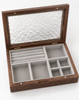 An open wooden jewelry organizer box with a textured glass lid, featuring multiple interior compartments lined in light gray velvet.