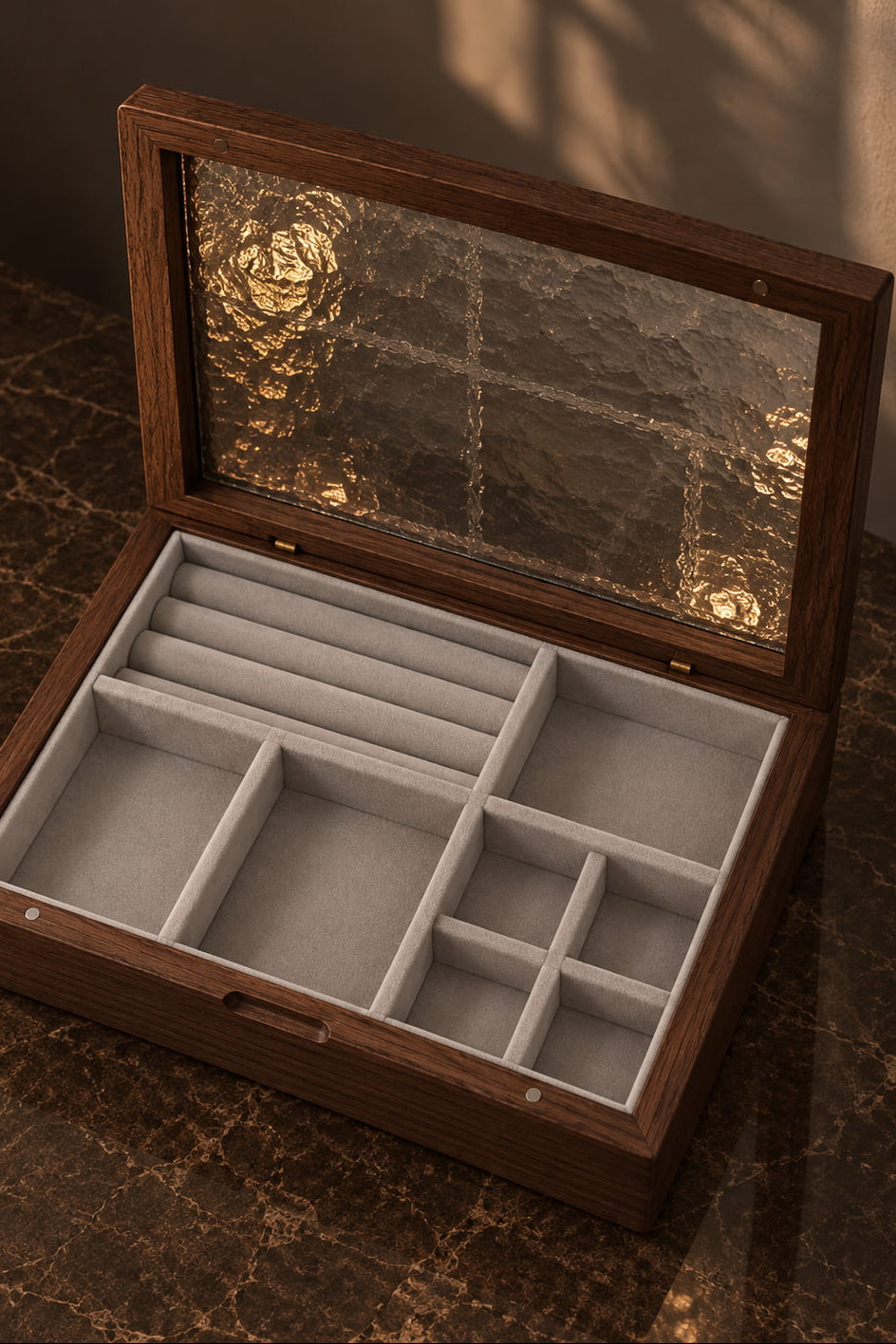 An open wooden jewelry organizer box with a textured glass lid, featuring light gray velvet interior compartments and ring rolls, set on a dark marble surface.